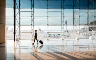 Matkustaja kävelee lentokentän terminaalissa matkalaukun kanssa, taustalla näkyvät lentokoneet korostavat modernia ja sujuvaa matkustuskokemusta.