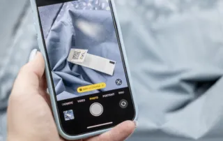 A person holding a smartphone displaying a photo of a textile label with a QR-code, placed on a blue garment. The phone screen shows a camera app in photo mode and a pop-up prompt to open the DPP platform.